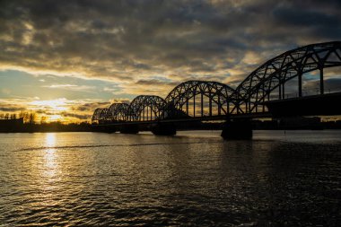 Gün batımında Riga 'daki Daugava Nehri üzerindeki ikonik kemerli çelik demiryolu köprüsü dramatik gökyüzünü, suyu ve akşam aydınlığına karşı tarihi ulaşım altyapısının silüetini gösteriyor.