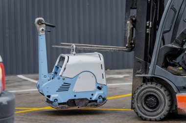 Endüstriyel bina yakınlarındaki beton yüzeyde forklift ile kaldırılan kompakt inşaat sıkıştırıcısı, ekipman taşımacılığı, lojistik operasyon ve ağır makinelerin güvenlik işlemleri