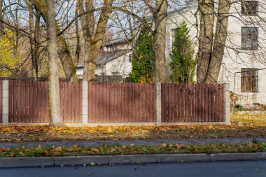 Kahverengi ahşap çit panelleri ve mimari dış tasarımı, sınır inşaatını ve banliyö manzarası organizasyonunu gösteren uzun ağaçlar arasında beton direkler bulunan yerleşim yeri.