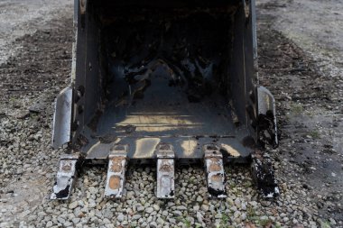 Sıkıştırılmış çakıl yüzeyine yerleştirilmiş kazı kovası yontulmuş kenarlar ve toprak kalıntısı, engebeli bölge koşullarını ve kazı araçlarındaki operasyonel stresi gösteren