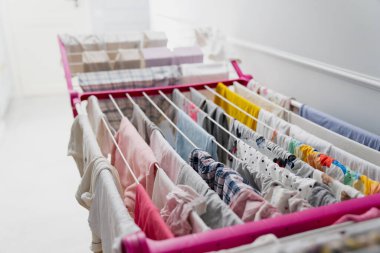 Temiz çamaşırlar, yukarıdan çekilen kapalı bir rafta birkaç kurutma hattına yayılmış durumda. Kumaş desenleri, renk iğneleri ve ev içi kurutma alanının düzenli yapısını vurguluyor.