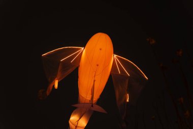 Kanat benzeri yarı şeffaf elementlerle aydınlatılmış turuncu hava heykel karanlık gece gökyüzüne karşı yüzüyor atmosferik sanatsal kurulum yaratıyor