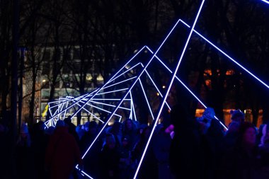 Parlak açılı mavi ışık çizgileriyle gece parkı kurulumu geometrik aydınlatılmış koridor oluşturarak hareket eden ziyaretçilerle dolu açık hava etkinliği atmosferi yaratıyor