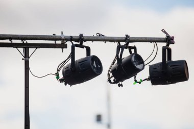 Açık hava aydınlatma teçhizatı. Üçlü sahne ışıkları metal destek çubuğunda asılı. Kablolama bağlantıları ve yapısal detaylar görünür.