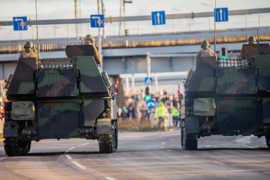 İki NATO zırhlı aracı, arka platformlarda görünen askerlerle bir şehir caddesi boyunca ilerliyor ve halk savunma gösterisi sırasında güzergahı izleyen izleyiciler var.