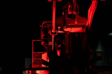 Düşük görünürlüklü teknik ya da endüstriyel ayarlar içinde kırmızı ışıkla güçlü şekilde aydınlatılan pistonları, kolları ve mekanik yapıları gösteren endüstriyel hidrolik bir makinenin ön yüzüne bakan perspektifi