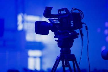 Profesyonel yayın kamerası bir tripoda monte edilmiş ve derin mavi olay aydınlatmasıyla aydınlatılmış, modern canlı üretim ortamlarında kullanılan teknik ekipmanları vurguluyor.