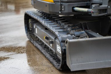 Islak asfalt üzerinde ön bıçak ve şasi parçaları olan endüstriyel kazı sistemi, inşaat makinelerinin çekiş ve istikrarını vurguluyor.