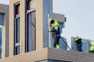 İnşaat işçisi çok katlı binalara cephe unsurları yerleştiriyor, modern inşaat endüstrisi uygulamaları, dış montaj çalışmaları ve kentsel kalkınma gösteriyor