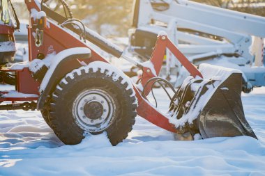 Taze karda dinlenen kompakt inşaat yükleyicisi, tekerleğin yakın görüntüsü, hidrolik kollar ve kovayla altın kış ışığı ve mekanik bahçe atmosferi