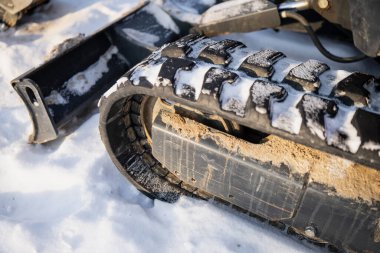 Kış boyunca, kar birikintisi, toprak lekeleri ve şasi paneli olan, parlak güneş ışığı altında endüstriyel ekipman içeren kauçuk sürüngen izlerini gösteren, takip edilmiş inşaat makinesinin yakın çekimi.