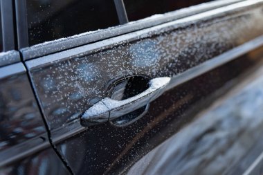 Aracın kapısının kışın kapandığı yerde buzlu kapı kolu ve dağınık buz noktaları, parlak siyah boya yansımaları ve bulanık sokak arkaplanı, soğuk hava ayrıntıları görülüyor.