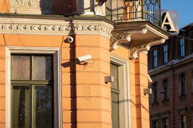 Sıcak turuncu dış görünüş dekoratif pervazlı balkon metal korkuluklu, yeşil pencere çerçeveli ve güvenlik kameralı şehir köşesinde parlak güneş ışığı.