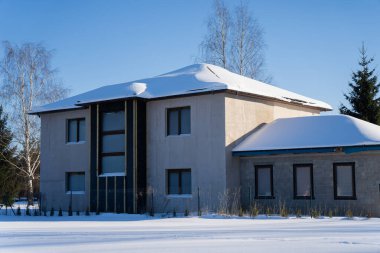 Geometrik cepheli ve geniş dikey camlı yeni bir aile evi, çatılarda kalın kar örtüsü, huş ağaçları ve temiz gökyüzü ile sakin bir kışlık arazi sahnesi.