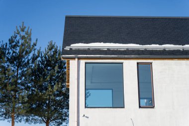 Minimalist ev dış görünüşü koyu çatı kiremitleri, saçaklarda kar, modern cam ve dikey pencere, açık kış gökyüzü ve çam ağacı siluetleri