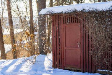 Karlı orman bahçesinde küçük kırmızı ahşap kulübe kapısı, karla kaplı çatısı ve duvarında kuru sarmaşıkları olan kırsal ek bina, kış güneşi ışığı ve çıplak ağaçlar.