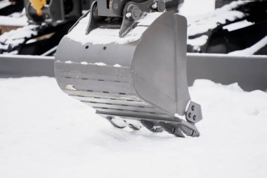 İnşaat kazısı kovası yakın görünüm, metal siper eklentisi aşınma izleri ve cıvatalar kenarda, karlı çalışma alanı yüzeyinin üstünde asılı.