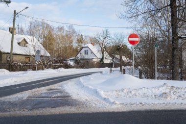 Kış kırsal yol eğrisi, giriş işareti yok, kar yığınları ve arka planda ağaçlar olan küçük evler, soğuk mevsim sürüş ve gündüz yol güvenliği kavramı
