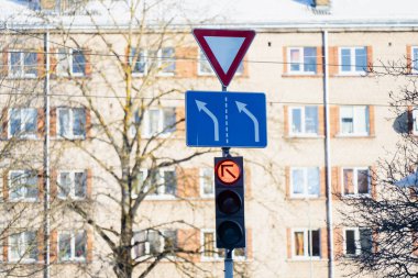 Mavi şeritli işaret levhası ve direğin üzerinde kırmızı trafik ışığı, apartman cephesi ve arka planda çıplak kış ağaçları