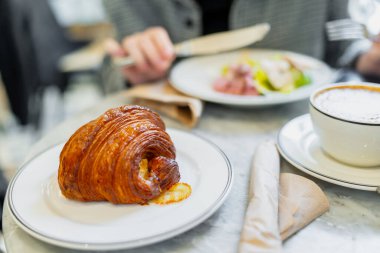 Sıcak kahvenin yanında servis edilen altın tereyağlı kruvasan tabakta yenen kişinin yumuşak tabağı, rahat kafe toplantısı ve hafta sonu kahvaltısı konsepti.