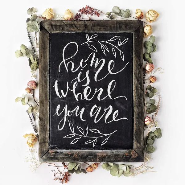 Phrase "Home is where you are" écrite en style calligraphie sur un tableau noir en bois avec des roses jaunes et roses et des feuilles vertes — Photo