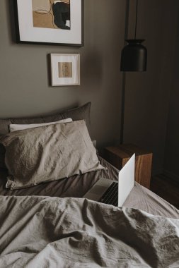 Modern iç tasarım konsepti. Hygge tarzı yatak odası, gri, yıkanmış çarşaf, yastık, dizüstü bilgisayar, ahşap komodin, asılı lamba. Minimalist İskandinav oturma odası.