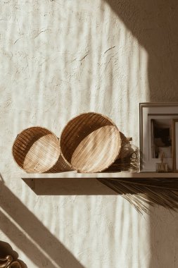 Modern boho tarzı ev dekorasyonu. Bohem dekorasyon aksesuarları: fotoğraf çerçeveleri, beton duvardaki ahşap raflarda saman plakalar. Sıcak güneş ışığı gölgeleri duvarda. Eski konsept.