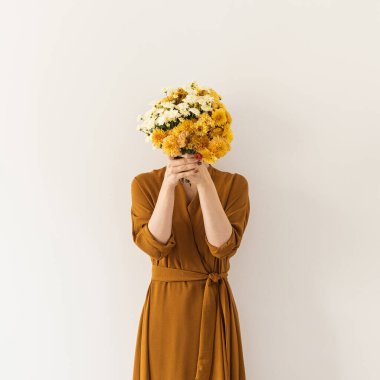 Kahverengi elbiseli genç bir kadın beyaz duvara bir buket kır çiçeği tutuyor. Asgari güzellik, moda konsepti. 