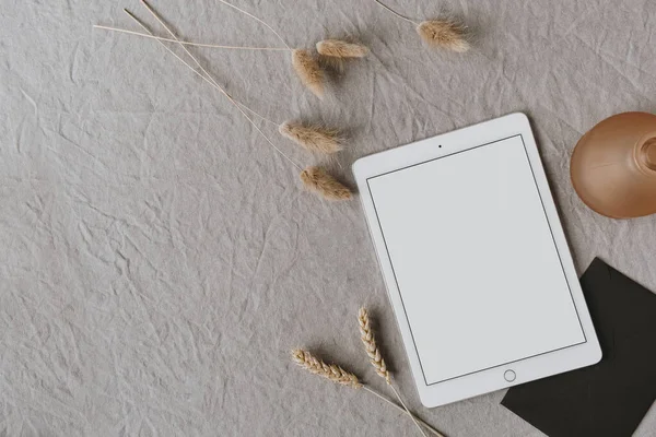 Boş tablet tabletlerin düzlüğü, pofuduk ponpon bitkileri, gri yıkanmış keten arka planda çavdar / buğday kulak sapları. Ofis masası çalışma alanı. İş, iş planı. Düz yatış, üst görünüm.