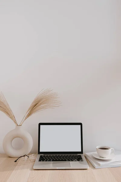 Female home office desk workspace. Blank screen laptop computer with copy space. Coffee cup, pampas grass in stylish vase on beige wooden table. Minimalist lifestyle blog mockup.