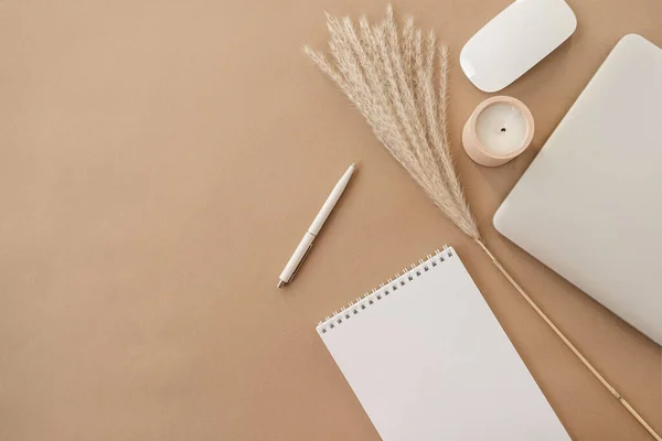 Flatlay of spiral flip notebook with blank paper sheet. Laptop, pampas grass, stationery on beige peachy pastel background table. Minimalist home office desk workspace. Top view mockup copy space.