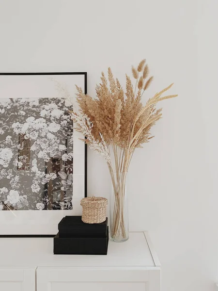 Modern nordic, scandinavian interior design concept. Beautiful floral frame, big vase with pampas grass and reeds, black boxes and straw casket on white commode. Minimal home concept.