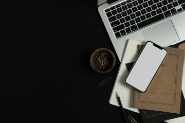 Boş ekran cep telefonu, dizüstü bilgisayar, defterler, siyah arka planda tahta kasede klipler. Ofis masası çalışma alanı. İş, iş planı. Düz yatış, üst görünüm.