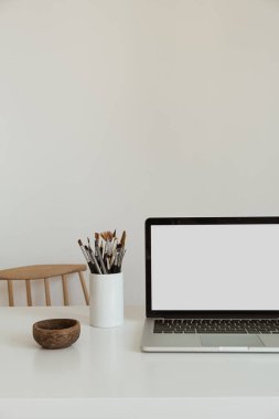 Masanın üzerinde beyaz duvara karşı kırtasiye malzemesi olan boş bir ekran olan dizüstü bilgisayar. Minimalist ev ofis çalışma alanı. Model şablonu.