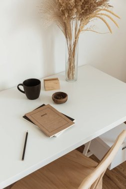 Estetik düşük ofis çalışma alanı iç tasarımı. Kupa, defter, pampa, çiçek buketi beyaz masada beyaz duvara karşı. Kız ve kadın patron ev işi konseptinde çalışıyor..