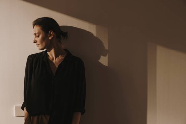 Duvarda güneş ışığı yansımaları var. Siyah gömlekli ve kahverengi pantolonlu güzel kadın duvarın önünde duruyor. Asgari estetik güzellik, dergi için moda konsepti, blog, sosyal medya.