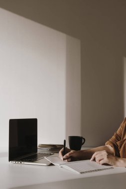 Kadın eli boş bir not defterine yazar. Minimalist ofis masası çalışma alanı. Masada kahve fincanı, kağıt çarşaf ve beyaz duvarda kırtasiye malzemesi olan dizüstü bilgisayar. İş, iş geçmişi.