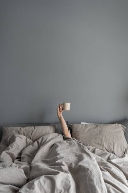 Kadın yatakta uzanıyor ve elinde kahveyle fincanı tutuyor. İyi sabahlar. Nötr İskandinav yatak odası, yatak odası, buruşuk çarşaf, gri duvarda yastıklar.