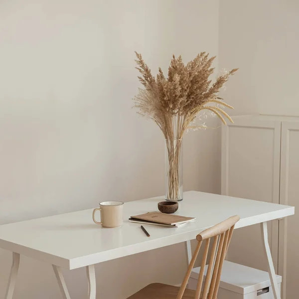 Women\'s home office workspace. Freelance work, business workplace. Light aesthetic hygge space with wooden chair, table, reed pampas grass bouquet, mug, notebook. Modern interior design.