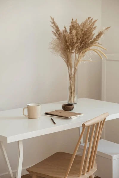 Women\'s home office workspace. Freelance work, business workplace. Light aesthetic hygge space with wooden chair, table, reed pampas grass bouquet, mug, notebook. Modern interior design.