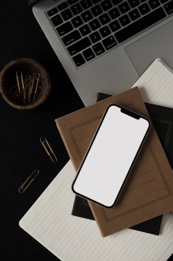 Boş ekran cep telefonu, dizüstü bilgisayar, defterler, siyah arka planda tahta kasede klipler. Ofis masası çalışma alanı. İş, iş planı. Düz yatış, üst görünüm.