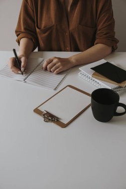 Kadın boş bir deftere yazıyor. Minimalist ofis masası çalışma alanı, kahve fincanı, dosya. İş, iş geçmişi.