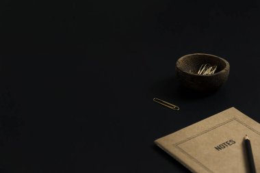 Kağıt kaplama defter, kalem, siyah arka planda tahta kasede ataçlar. Minimalist estetik ofis masası çalışma alanı. İş, iş kavramı.
