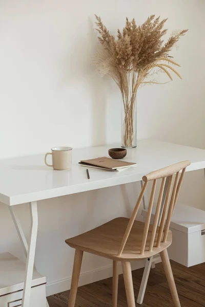 Aesthetic minimal office workspace interior design. Mug, notebook, pampas grass floral bouquet on white table against white wall. Girl, woman boss work at home business concept.