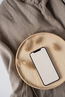Boş ekran cep telefonu, ahşap tepside tavşan kuyruğu çimeni ve buruşuk keten kumaş battaniyesi. Düz yatak, üst görünüm minimalist blog, web sitesi, uygulama şablonu. Boşluk modellemesini kopyala.