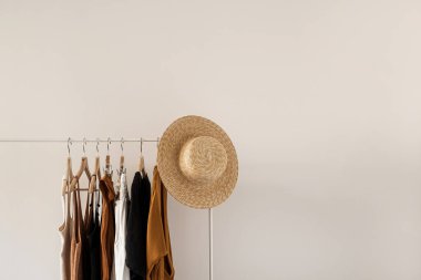 Estetik minimalist moda etkileyicisi blog kompozisyonu. Süslü pastel yaz kadın kıyafetleri, elbiseler, üstler, tişörtler, beyaz duvara asılı elbise askısı üzerindeki hasır şapka. Moda kadınları