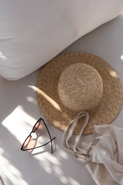 Kadın moda aksesuarları. Şık kadın gözlükleri, hasır şapka, beyaz kanepede yastıklı alışveriş torbası. Moda blogu, web, sosyal medya için yaratıcı kompozisyon. Üst görünüm