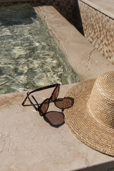 Sunglasses and straw hat on marble swimming pool side with clear blue water with waves sunlight shadow reflections. Minimal aesthetic summer vacation concept background.