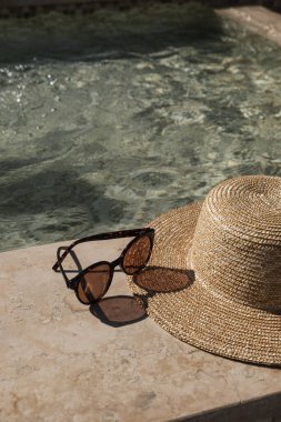 Güneş gözlüğü ve hasır şapka mermer yüzme havuzu kenarında açık mavi su ve güneş ışığı yansımaları. Minimum moda estetik yaz tatili yaratıcı geçmişi
