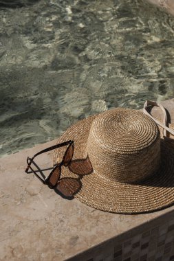 Güneş gözlüğü ve hasır şapka mermer yüzme havuzu kenarında açık mavi su ve güneş ışığı yansımaları. Minimum estetik yaz tatili konsepti.